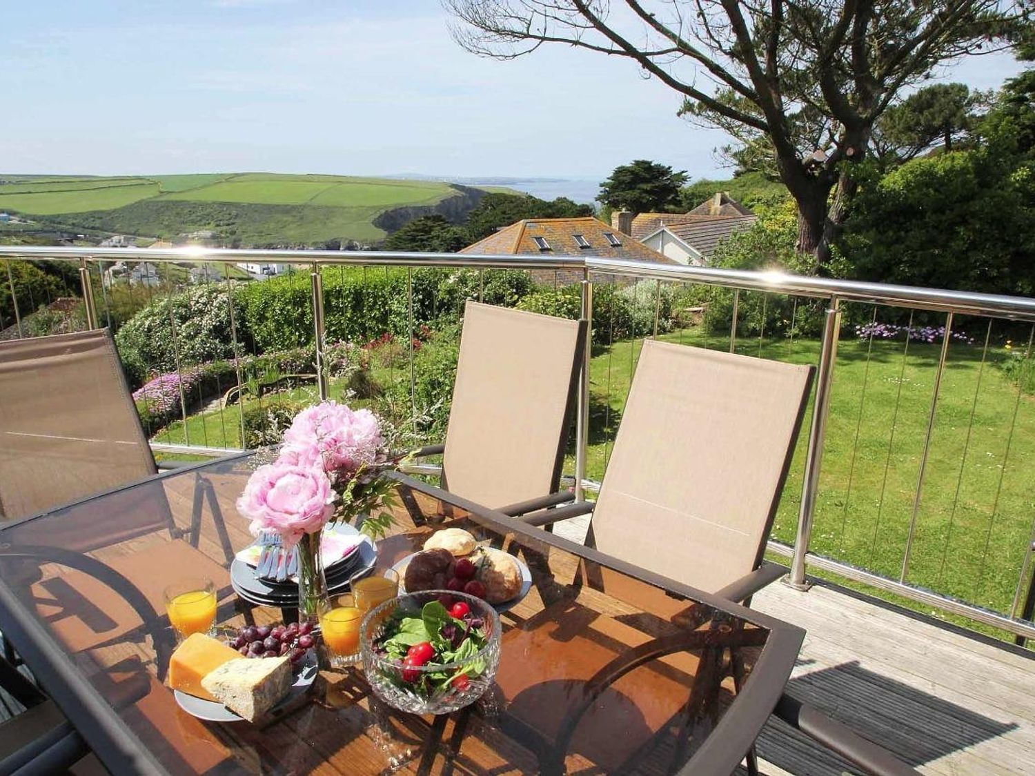 An outdoor dining table with food and drinks on a balcony overlooking a garden and green hills at Windrush in Mawgan Porth