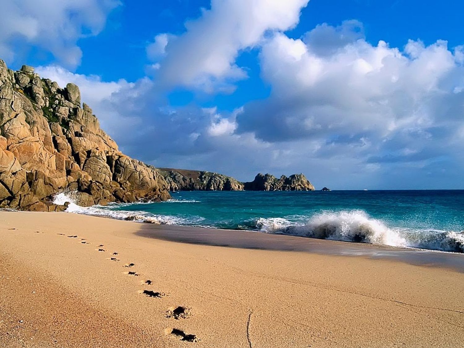 A sandy beach with footprints leading towards rocky cliffs and waves at Porthcurno Bay View in Porthcurno