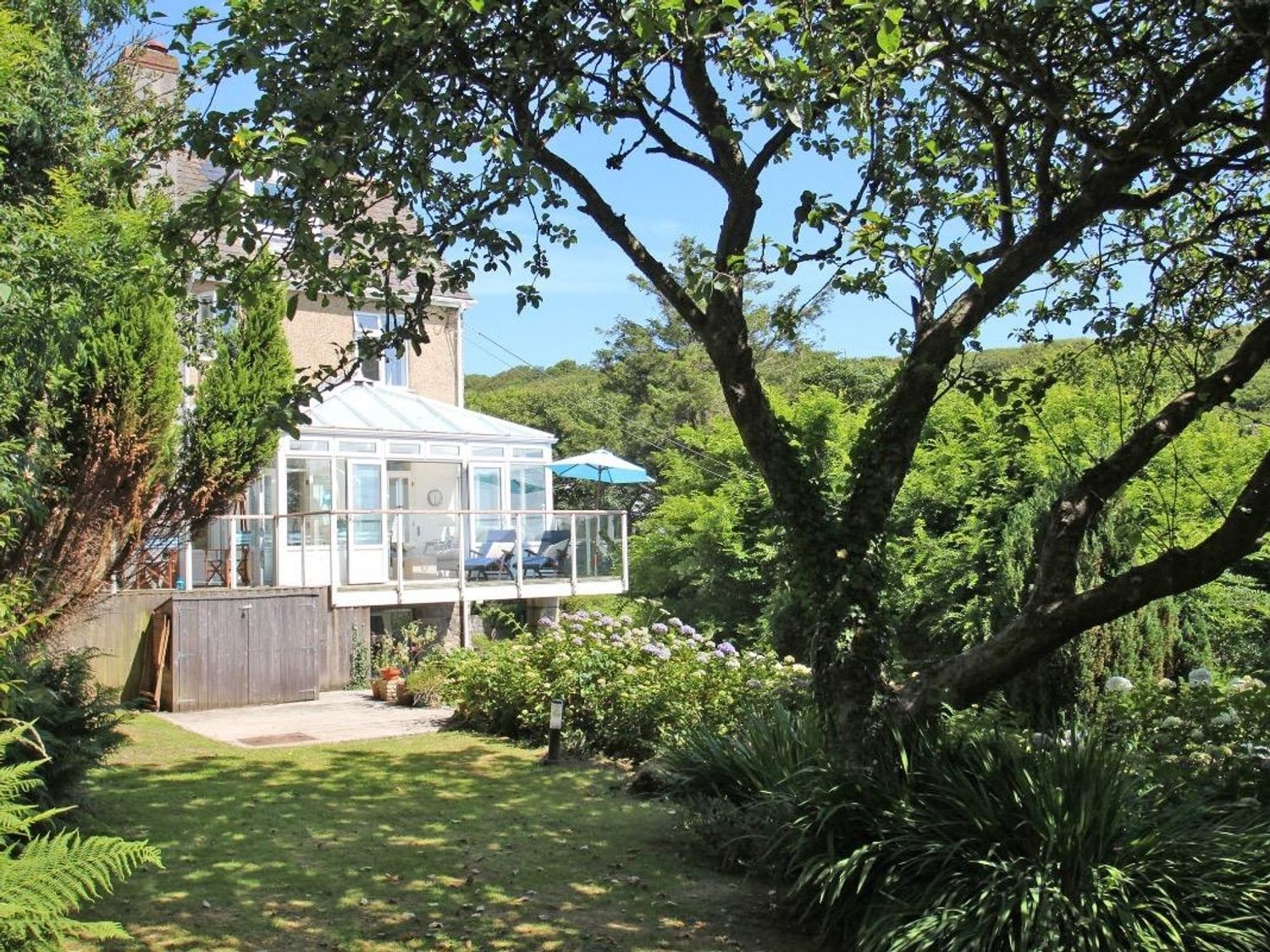 A garden with trees and bushes near a house with a glass conservatory and patio furniture at Porthcurno Bay View in Porthcurno