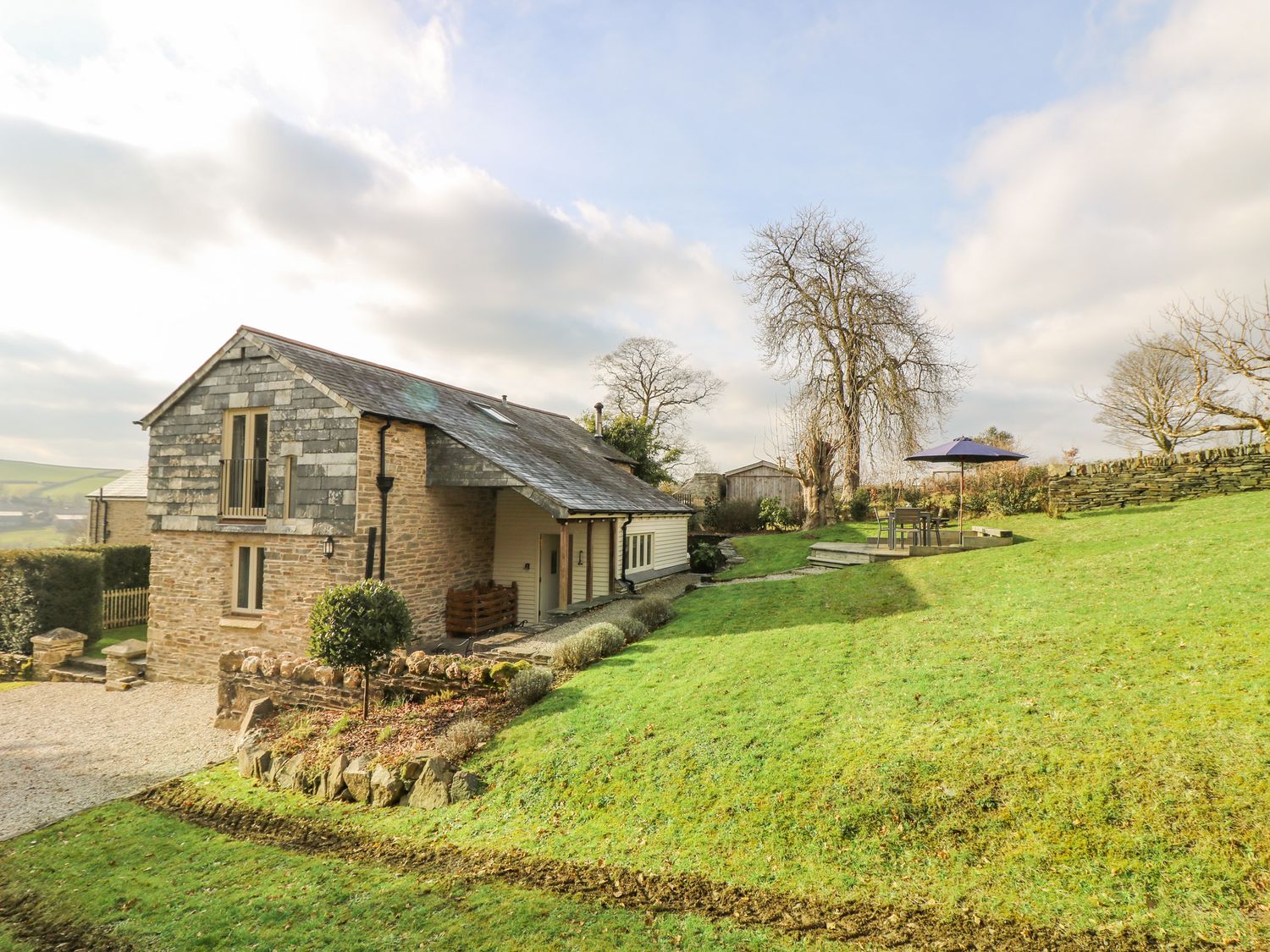 A stone and wood house with a slate roof on a grassy hill with outdoor seating and umbrella at Woodfield Coach House in Merrymeet