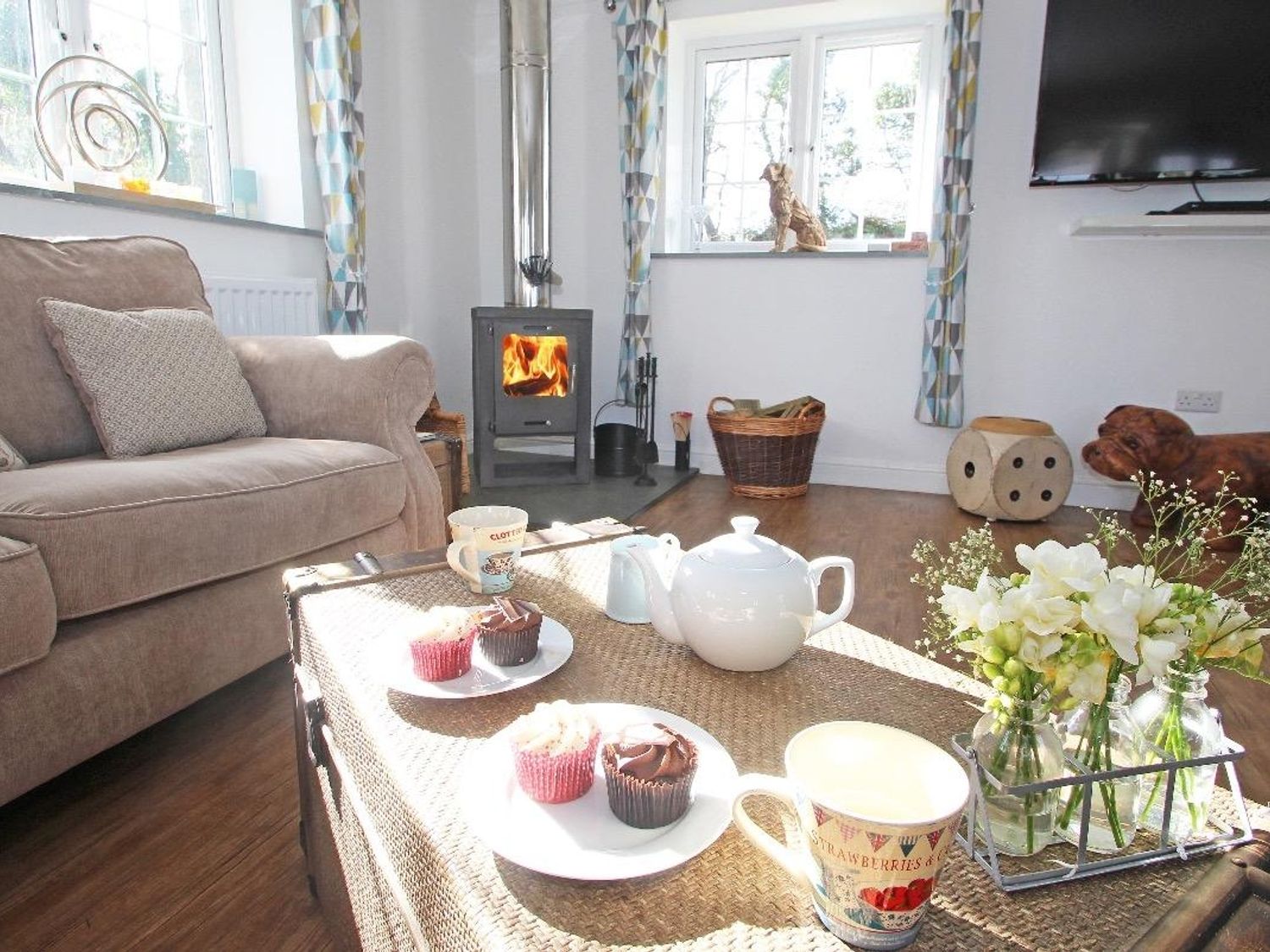 A living room with a sofa a wood burning stove a coffee table with teapot cups cupcakes and flowers at Omalast in Chacewater near Truro