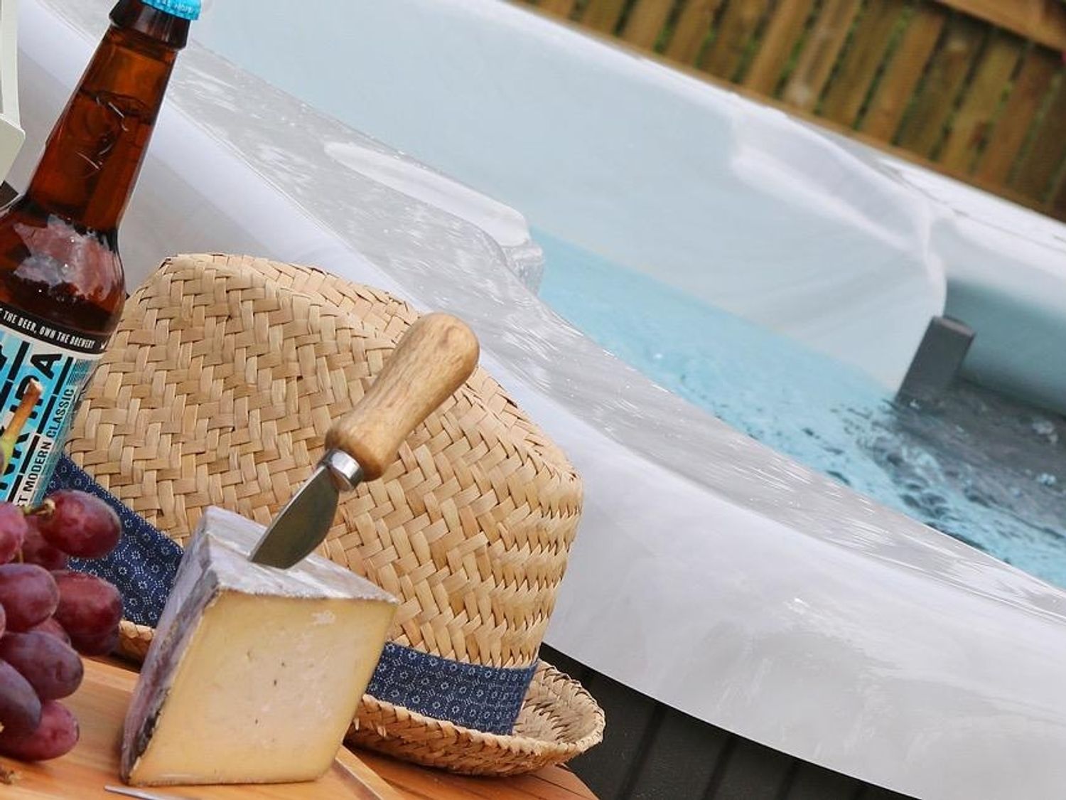 A close-up of cheese with a knife, a bunch of grapes, a bottle of beer, and a straw hat next to a hot tub at Omalast in Chacewater near Truro