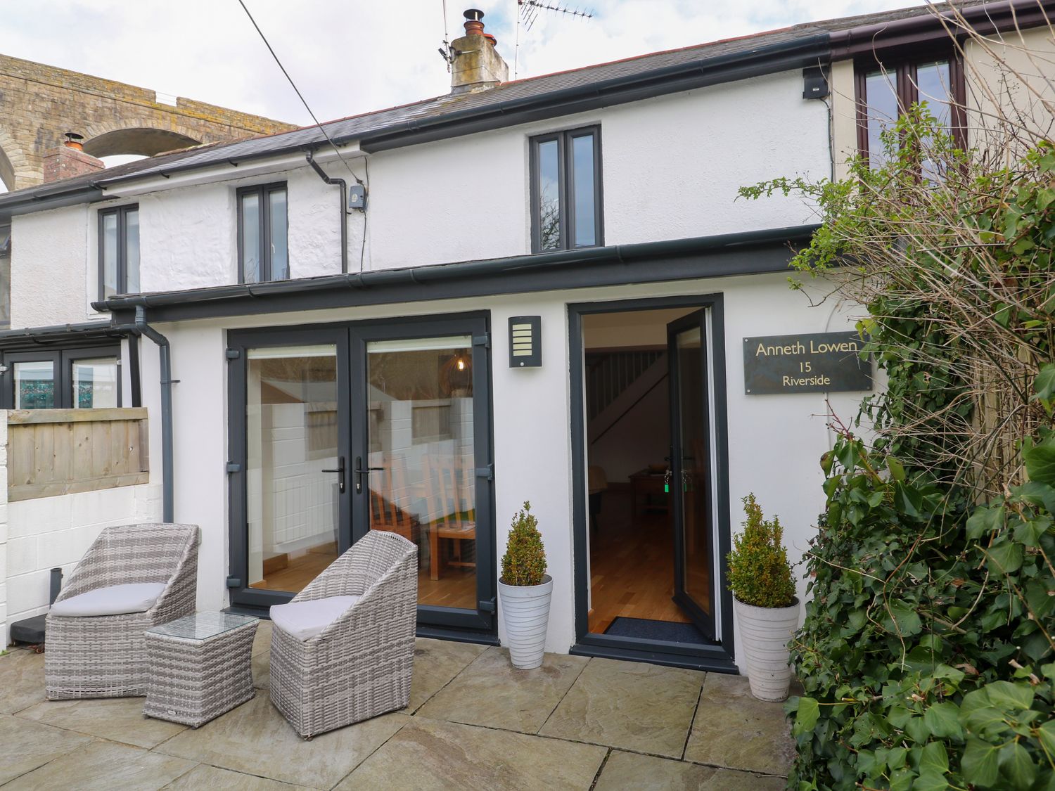 A patio with two wicker chairs and a small table outside a white house with dark framed doors and windows at Anneth Lowen Angarrack near Hayle