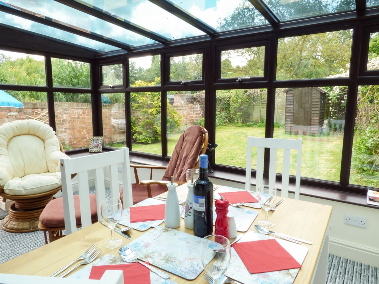 A dining table set with glasses utensils and red napkins in a conservatory with chairs and a garden view at 27 Swan Street in Boxford