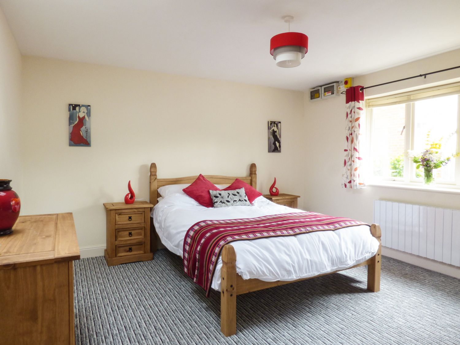 A bedroom with a wooden bed white bedding red pillows and blanket two wooden nightstands with red decorations wooden dresser and window with floral curtains at 27 Swan Street in Boxford