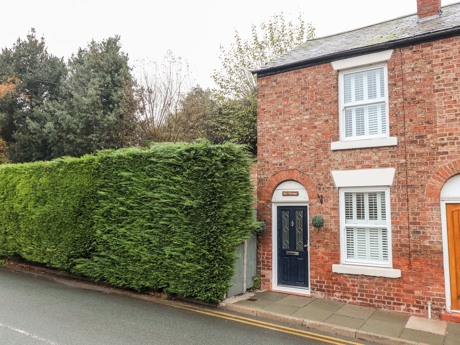 A brick house with a dark blue door and white windows next to a tall green hedge at Elv Cottage in Chester