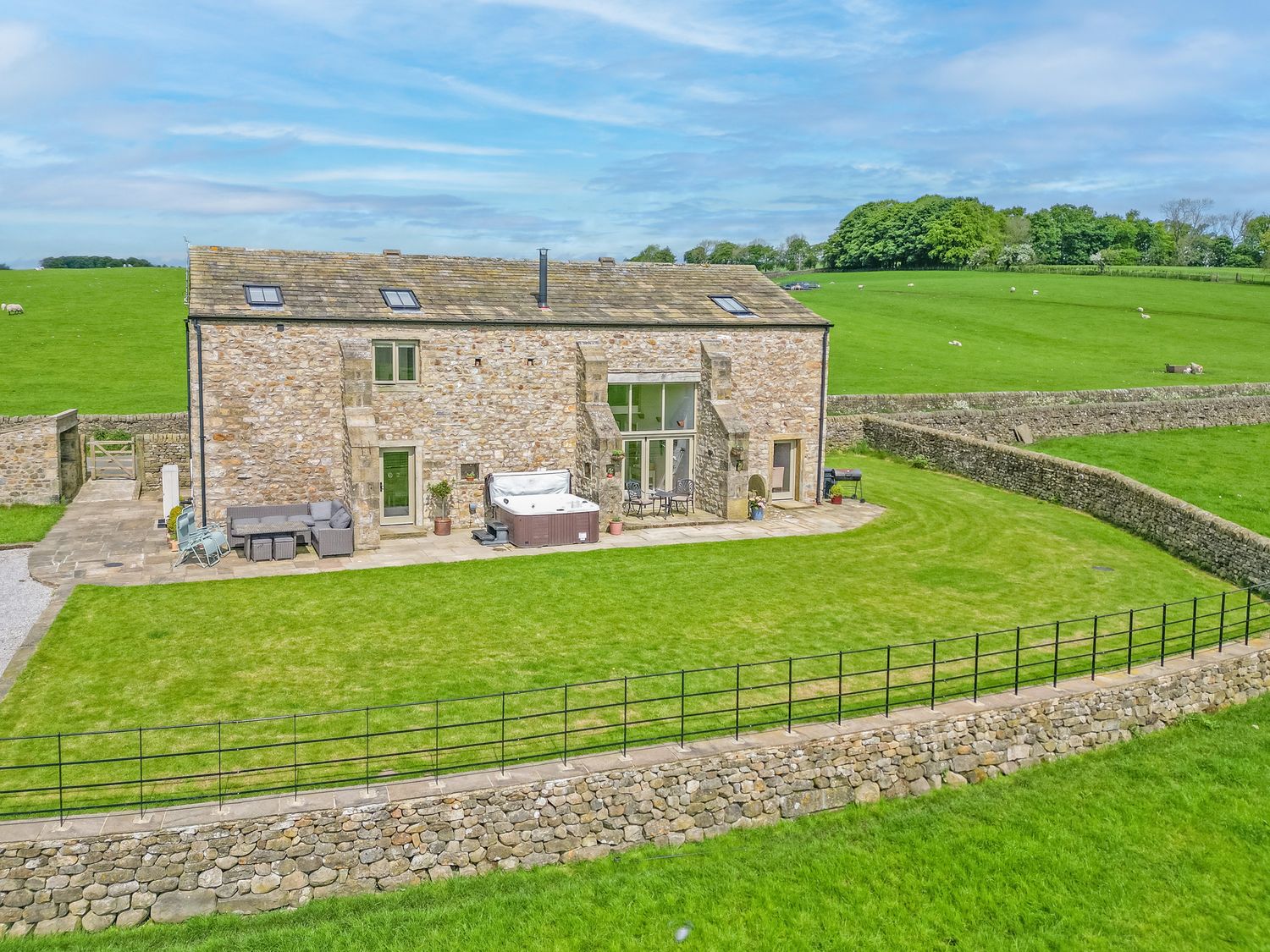 A stone-built house with a patio garden lawn outdoor seating and hot tub surrounded by stone walls in a rural field at Crane Field Laithe in Otterburn North Yorkshire
