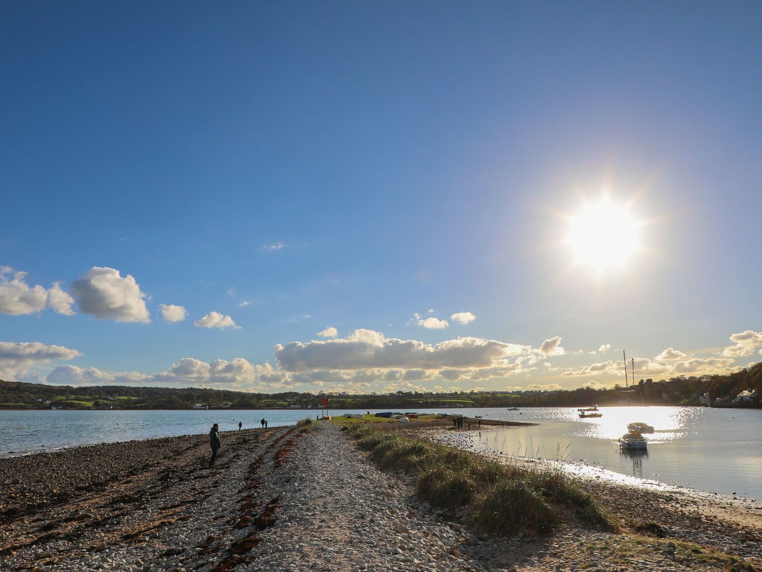 A rocky shoreline with a few people walking on the beach and boats anchored in the water at Glan Dwr in Red Wharf Bay