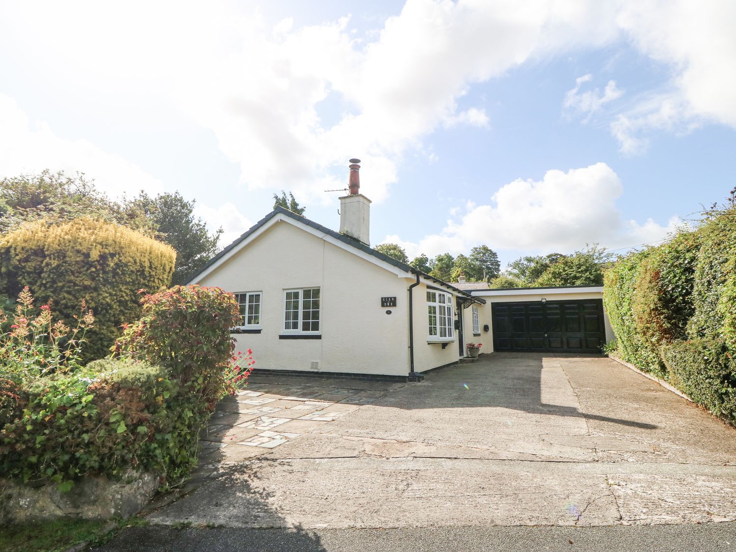A single story house with a driveway and black garage doors surrounded by hedges and bushes at Glan Dwr in Red Wharf Bay