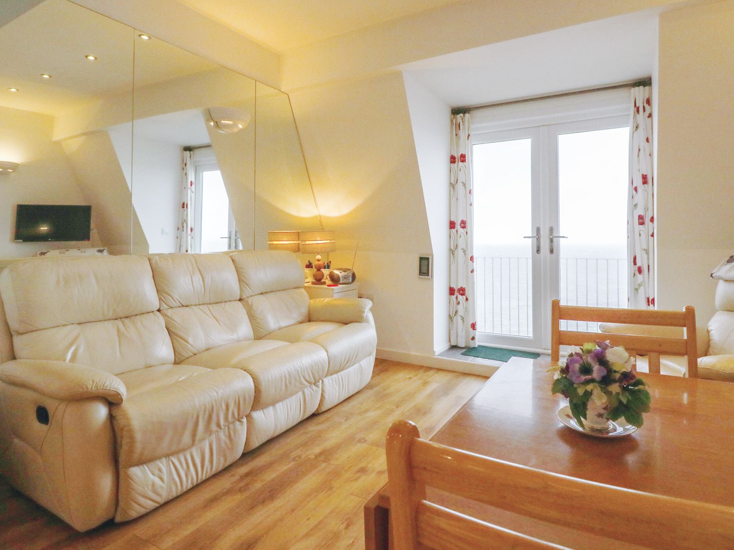 A living room with a beige leather sofa a wooden table with flowers and glass doors with floral curtains at 10 Oceanside in Ilfracombe