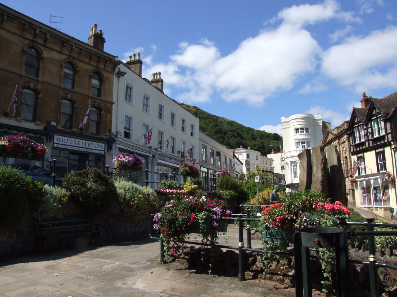 A street with flower arrangements and shops including Malvern Curtains in Malvern