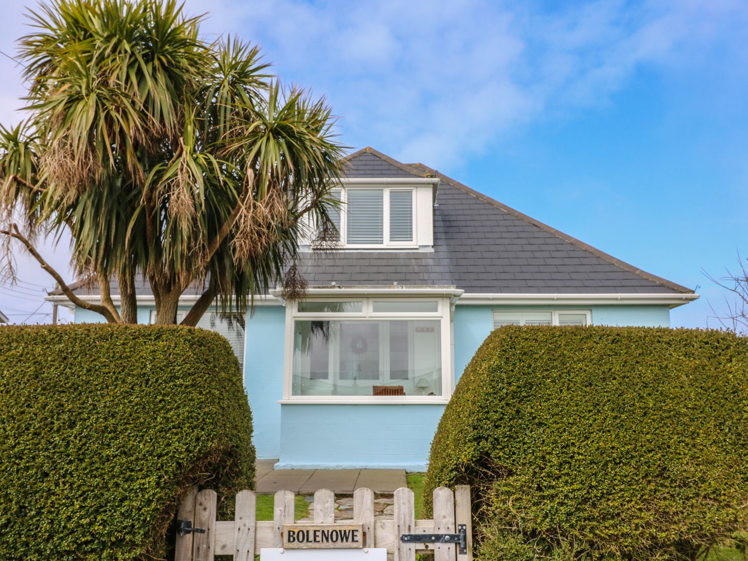 A house with blue walls and a gray roof behind trimmed hedges and a wooden gate with a sign Bolenowe in Trebetherick near Polzeath