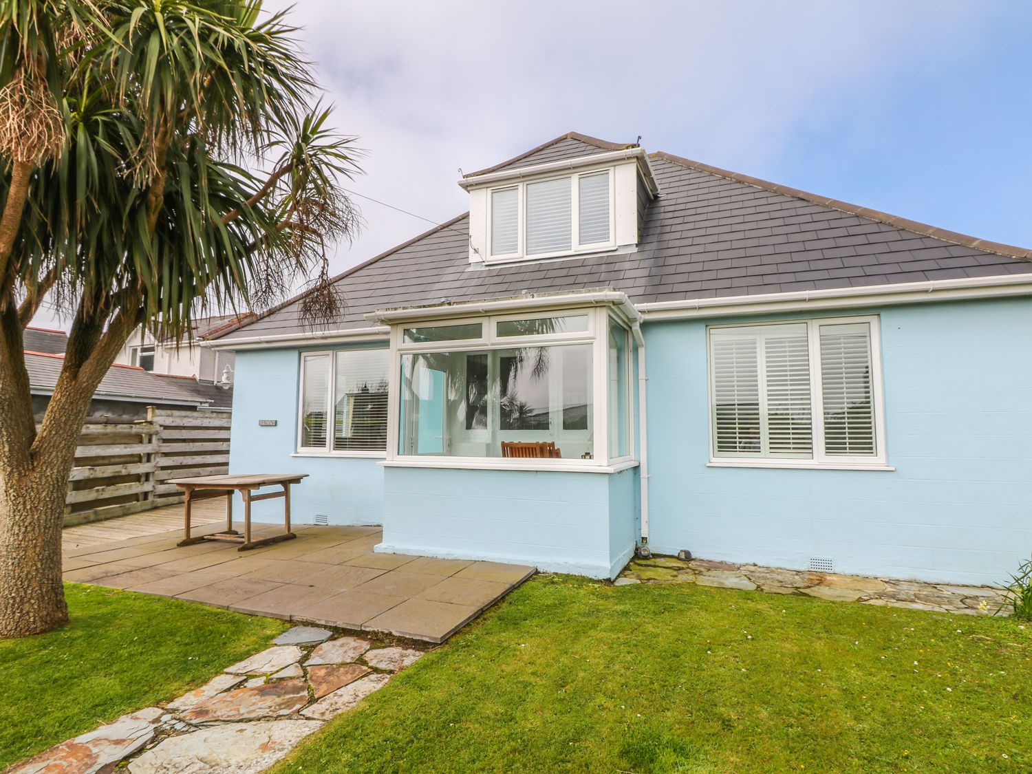 A blue house with a tiled roof and white-framed windows a wooden table patio area a palm tree and a stone pathway in the garden at Bolenowe Trebetherick near Polzeath