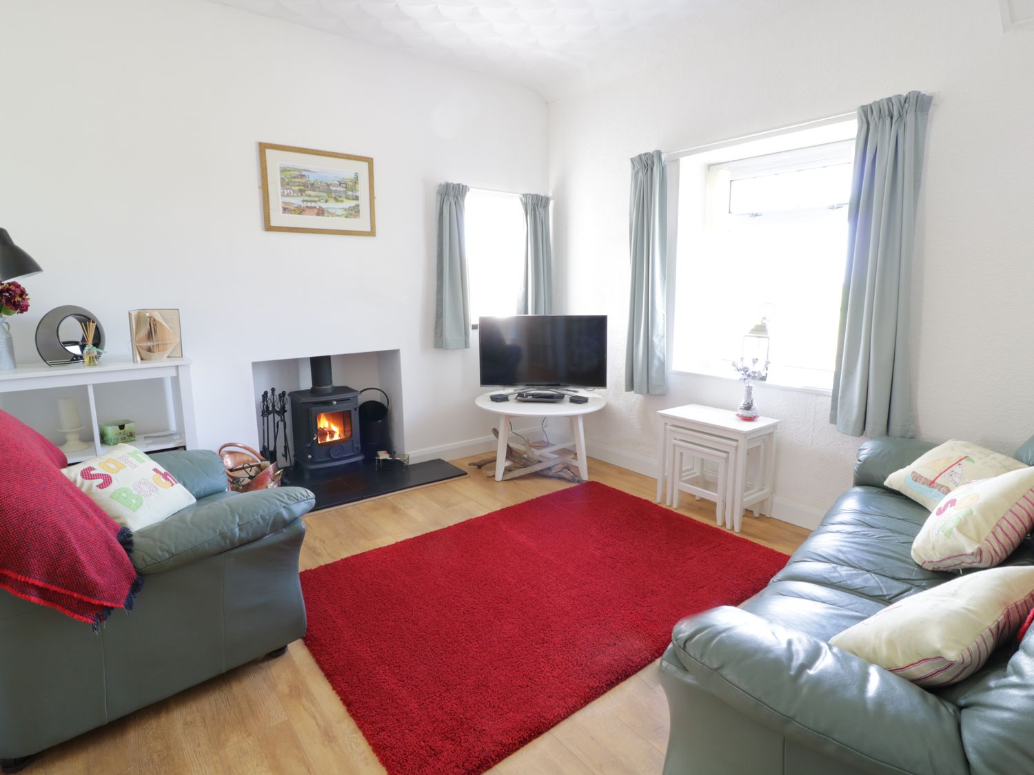 A living room with green sofas a red rug a wood burning stove and a television on a white table at Trigfa Cottage in Abersoch