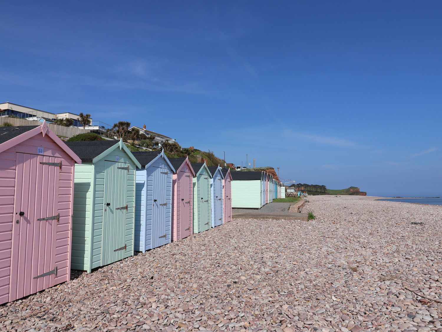 A row of pastel-colored beach huts on a pebble beach with houses on a hill in the background at Robyn Cottage in Otterton