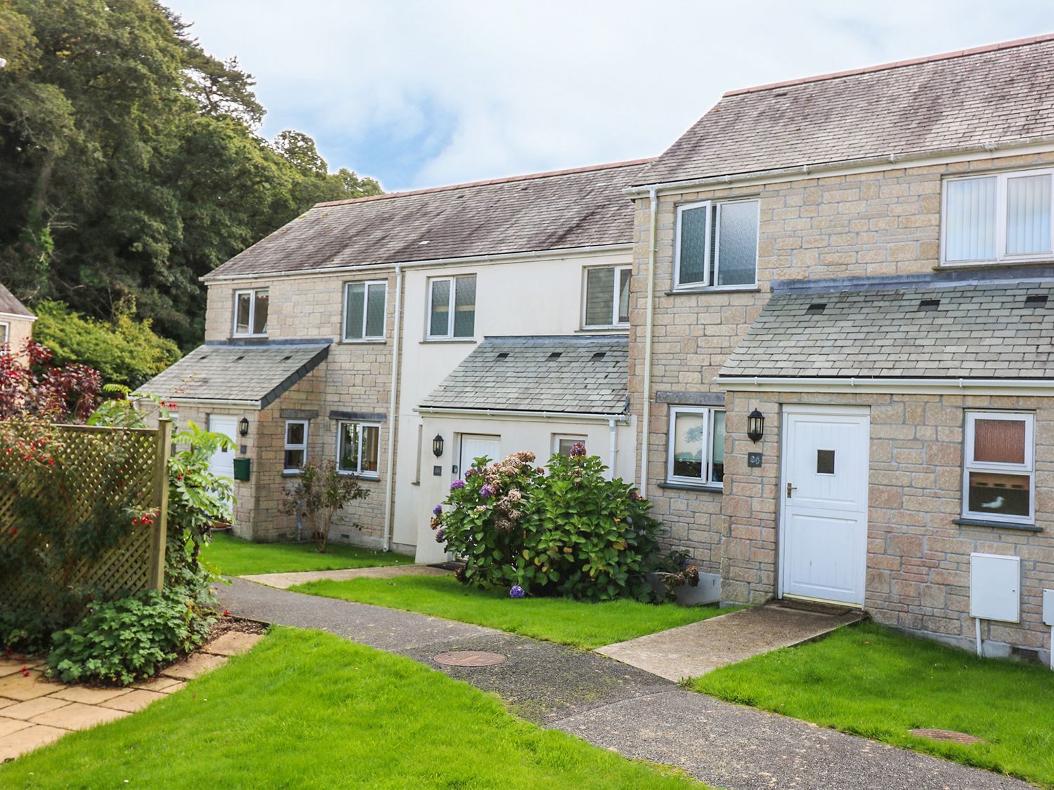 A row of stone and brick terraced houses with white doors and windows along a paved path with green lawns and shrubs Oyster Cottage in Falmouth