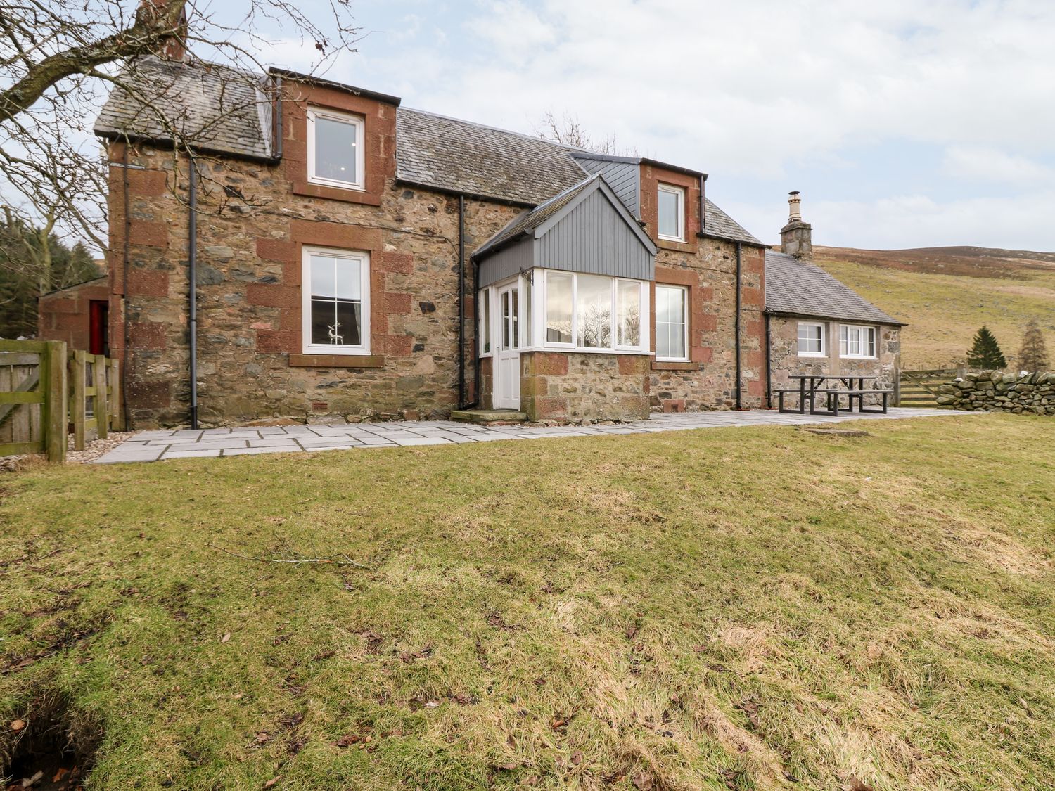 An exterior view of a stone house with multiple windows and a small porch on a grassy area at White Hillocks Farm House in Kirriemuir