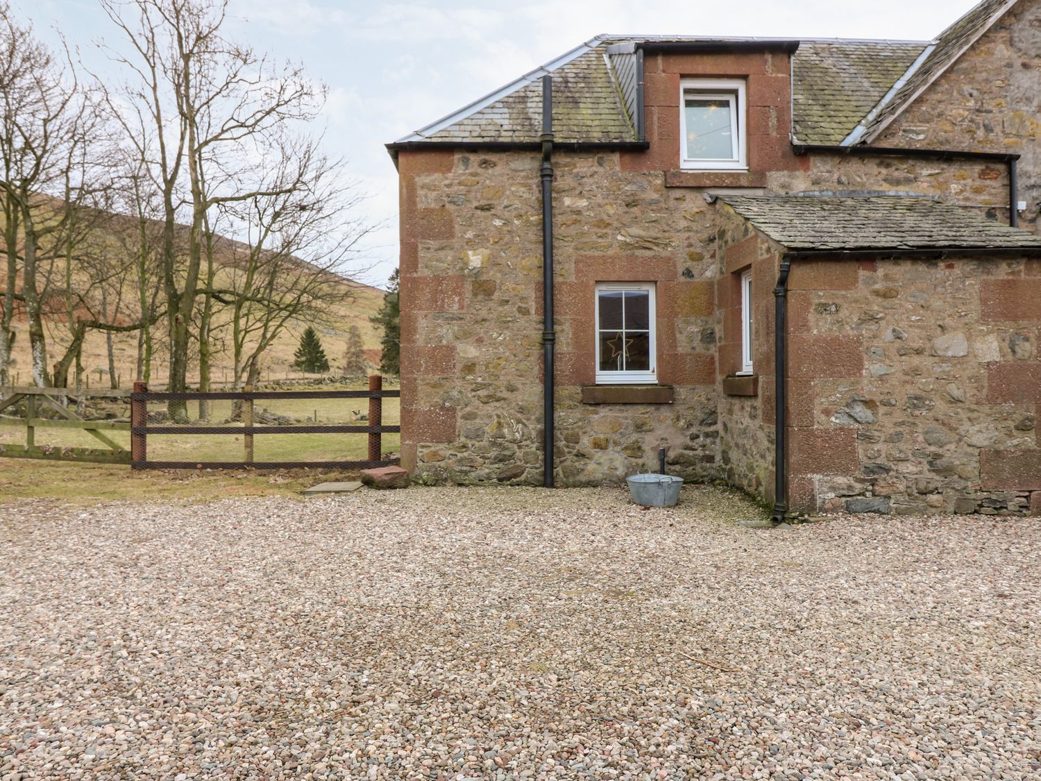 Stone house with gravel yard and wooden gate with trees and hills in the background at White Hillocks Farm House Kirriemuir