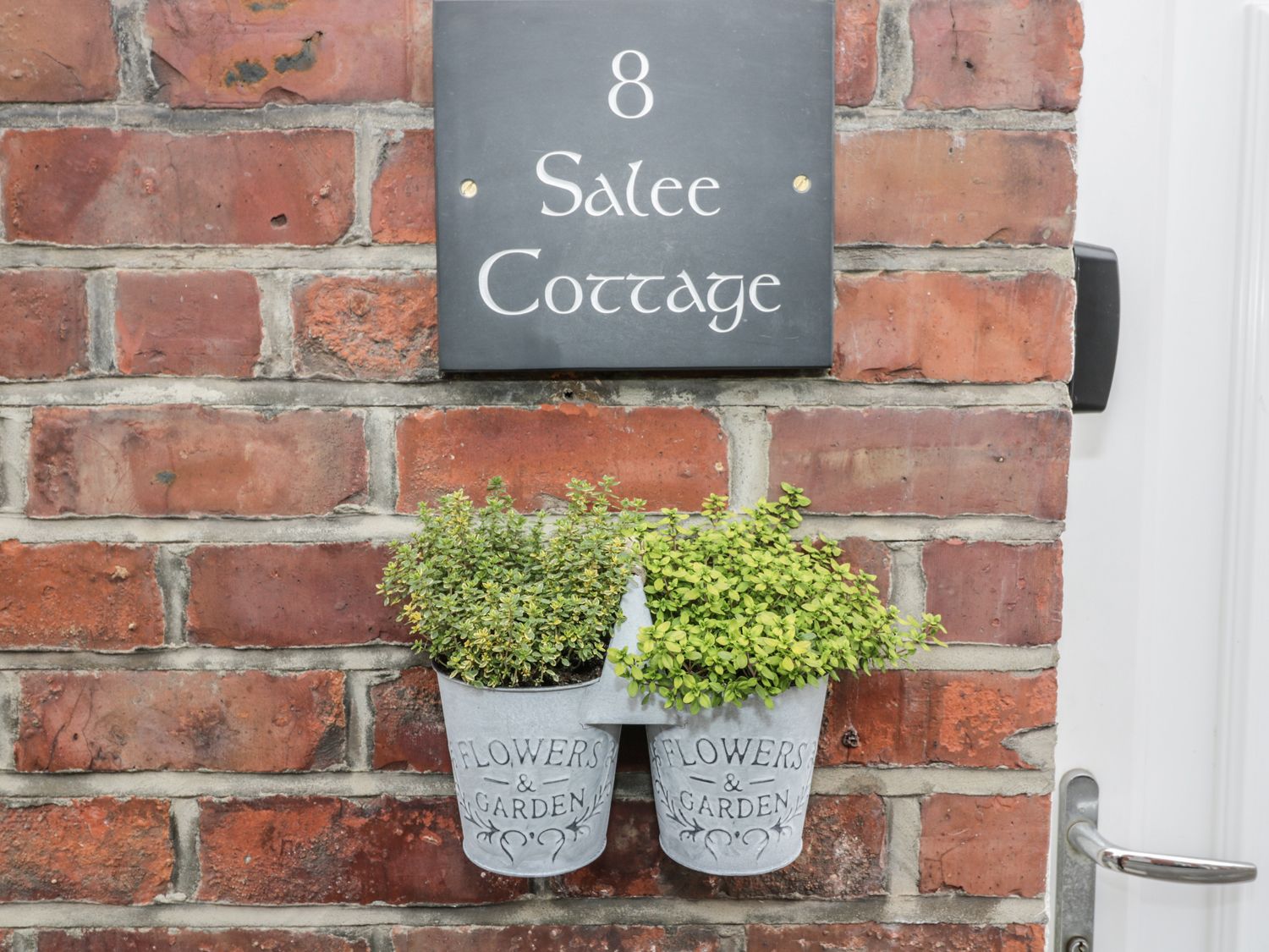 A brick wall with a sign reading 8 Salee Cottage and two metal pots with plants near a door handle at Salee Cottage in Newbiggin-by-the-Sea