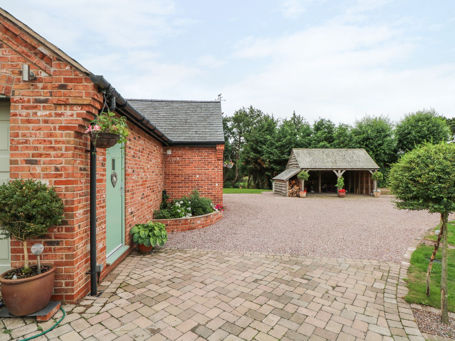 A brick building with a green door and potted plants beside a gravel driveway and a wooden carport surrounded by trees at Well View Cottage in Chester