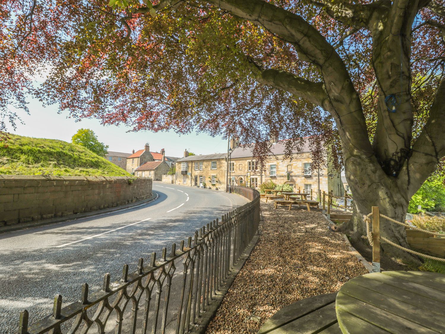 A curved road with stone buildings and picnic tables under a large tree at The Olde Stables in Warkworth