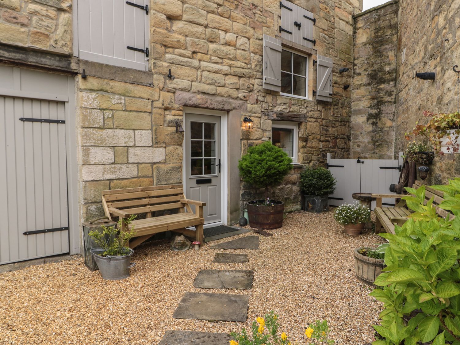 An outdoor courtyard with stone walls wooden benches potted plants and a gravel walkway at The Olde Stables in Warkworth
