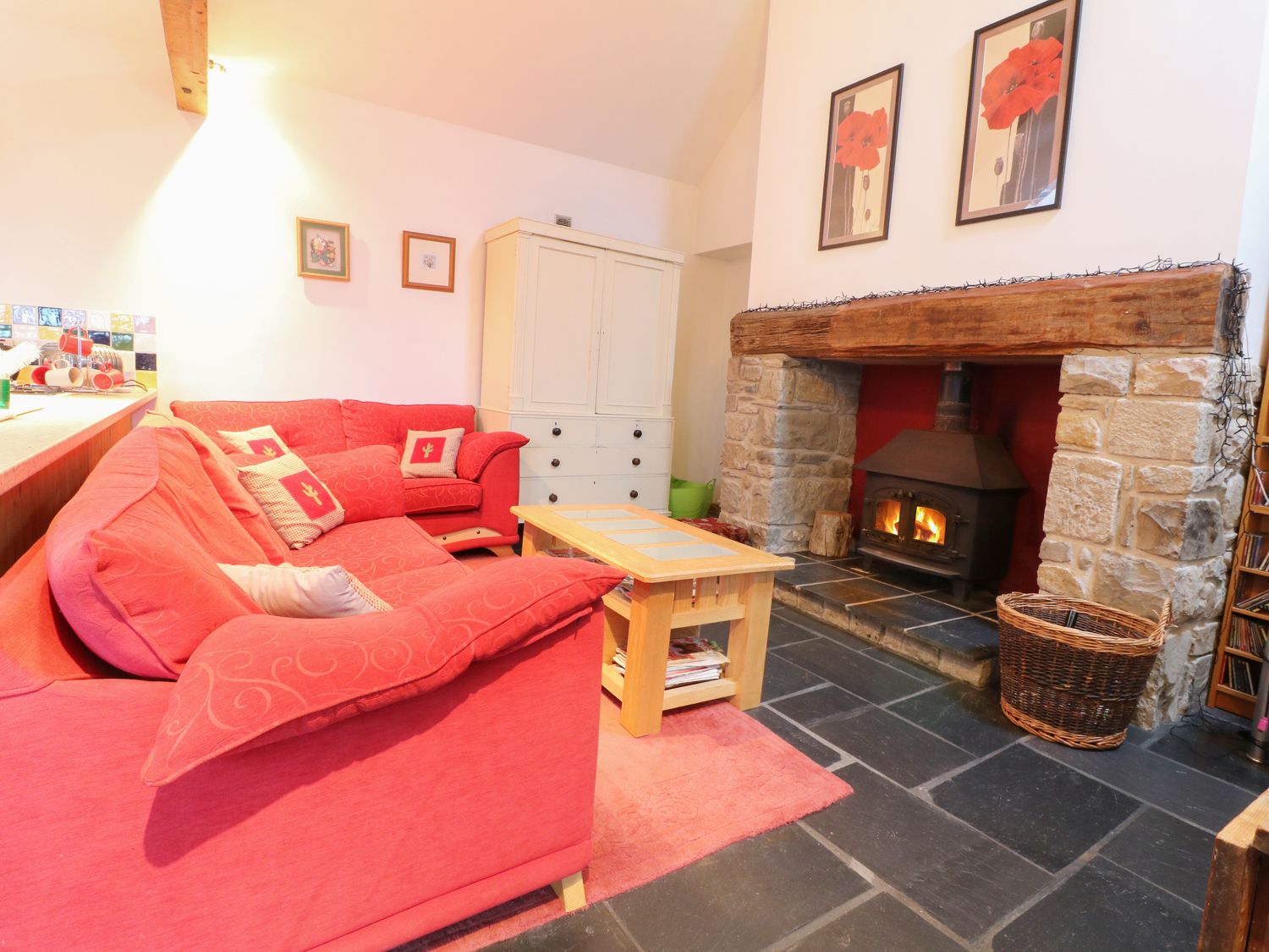 A living room with red sofas a wooden coffee table a stone fireplace with a wood stove and framed flower pictures on the wall at Beudy Mawr in Y Felinheli