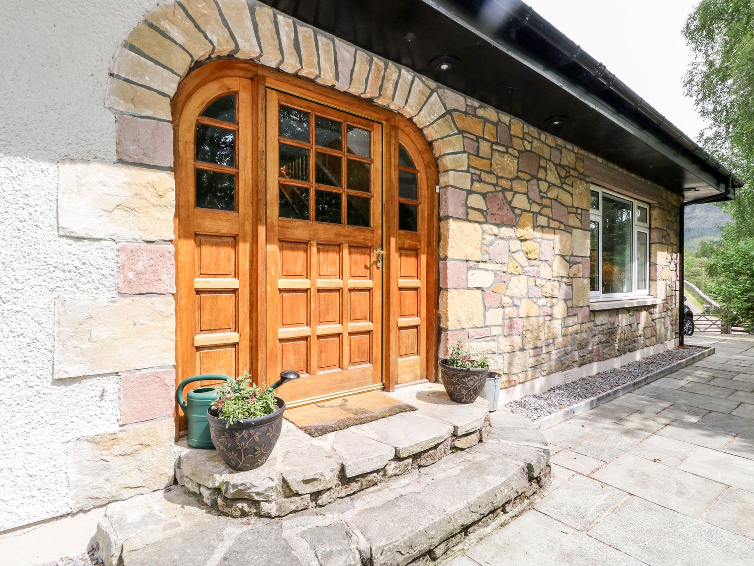 A wooden arched front door with glass panels set in a stone wall with two plant pots and a watering can at Tigh Na Drochit in Garve