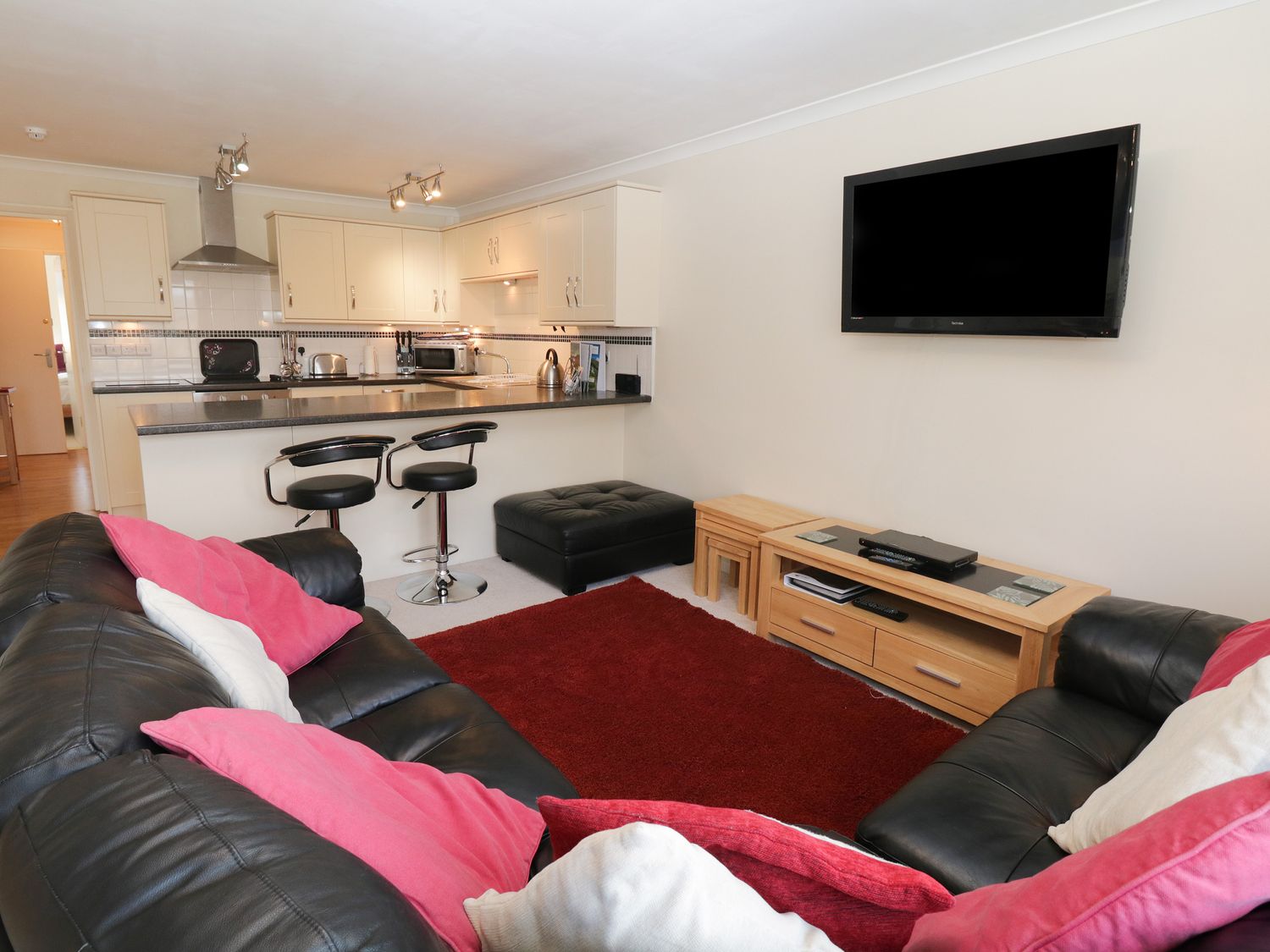 A living room with black leather sofas and a red rug facing a wall-mounted TV and an open kitchen with a breakfast bar and stools at Cumbria Court in Windermere