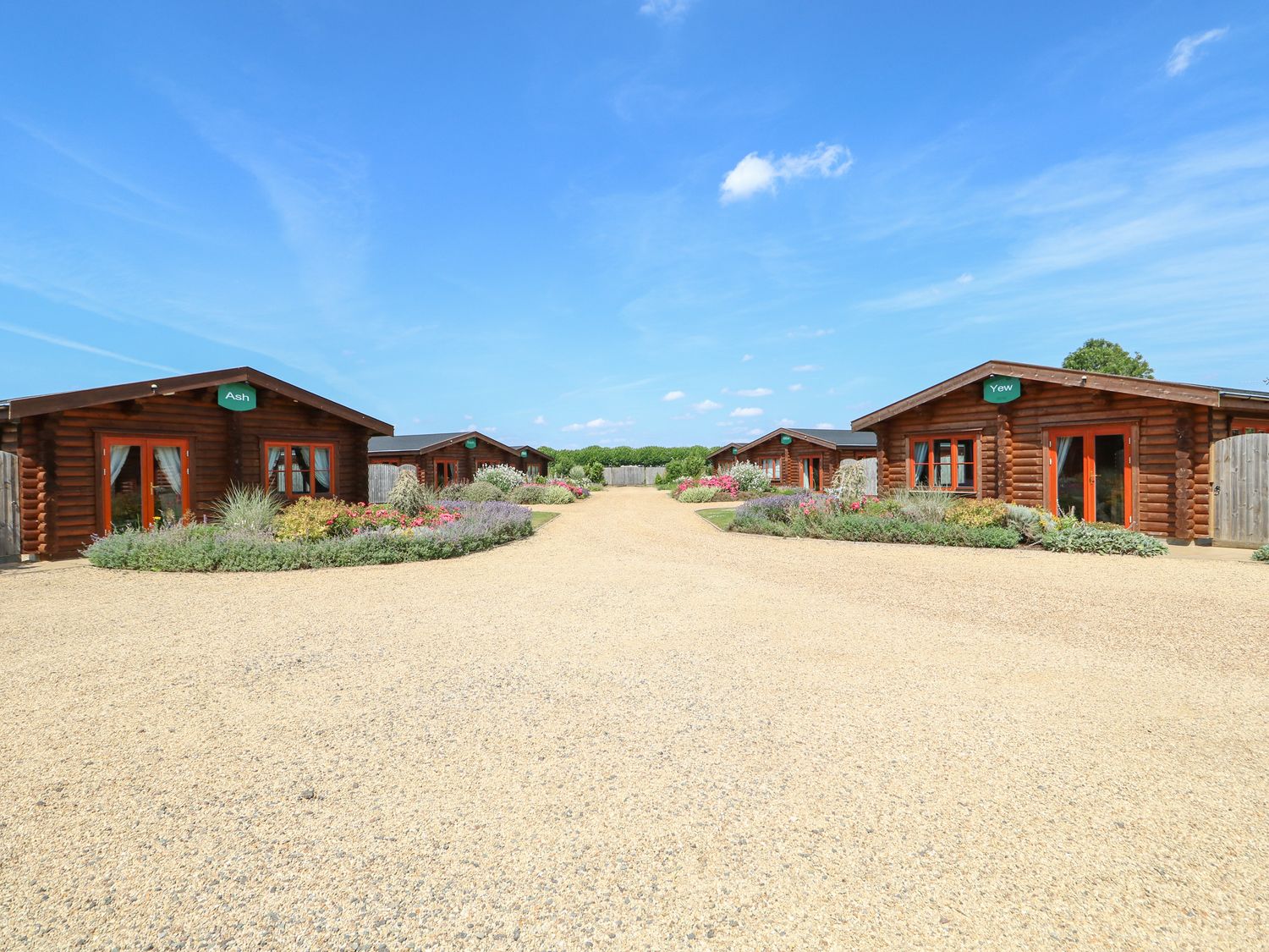 Four wooden lodges with gravel driveway and garden beds under a blue sky at Ash Lodge in Greetham Rutland