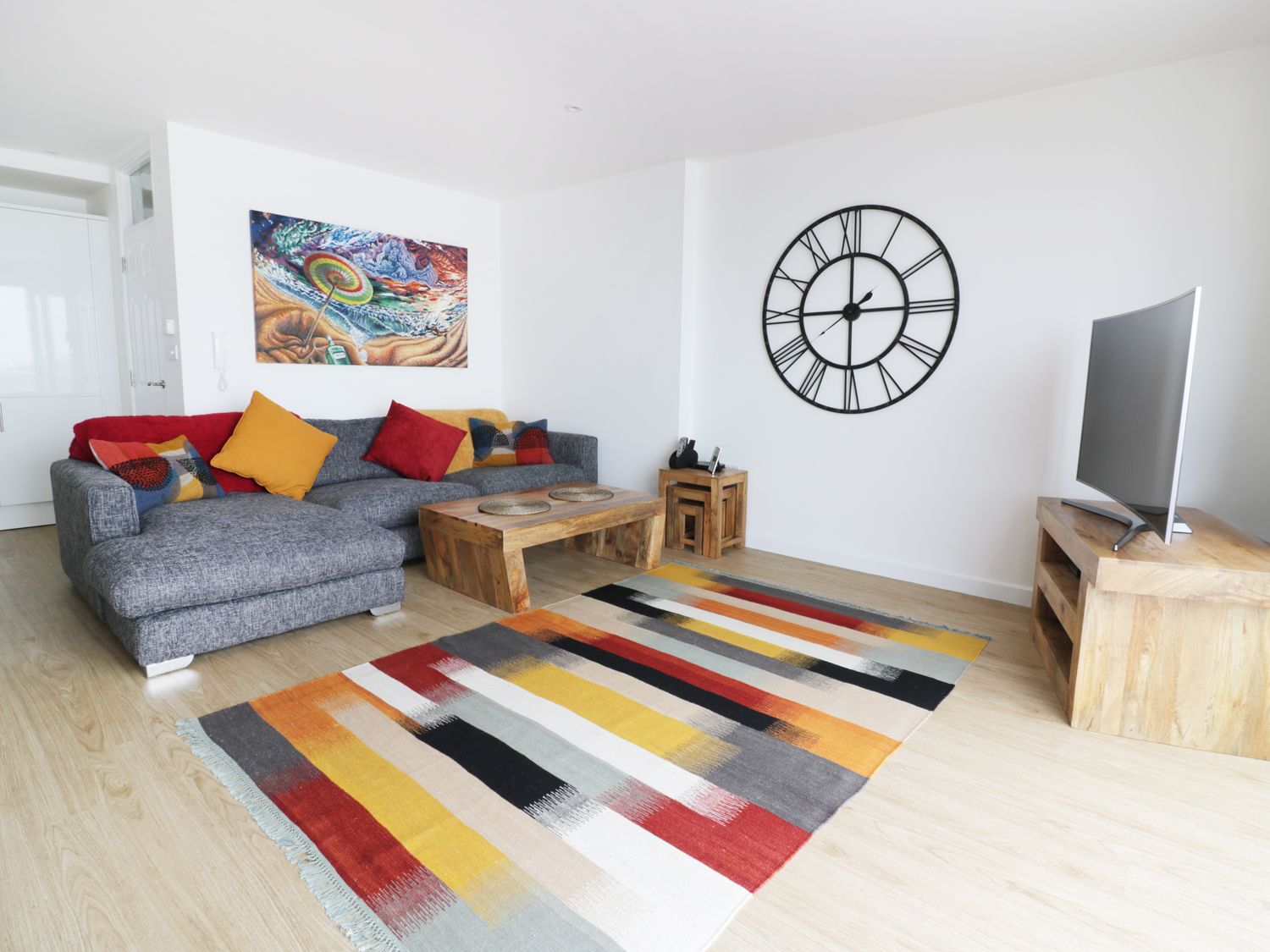 A living room with a gray sofa with colorful cushions a wooden coffee table a large wall clock a flat screen tv on a wooden stand and a colorful striped rug at The View in Newquay