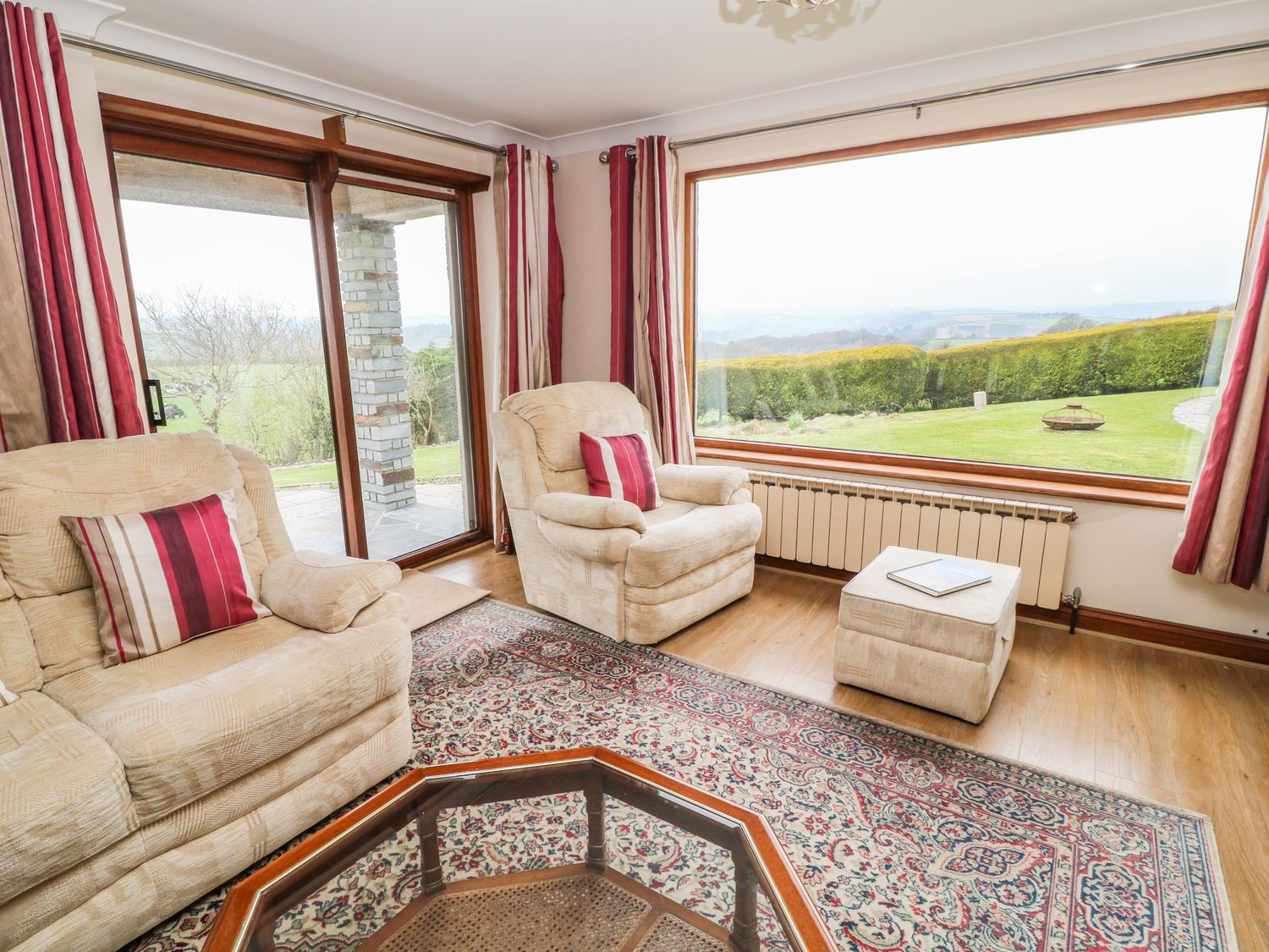 A living room with beige armchairs and a sofa with red and white cushions a glass coffee table a patterned rug large windows overlooking a garden at Higher Kernick Farm in Launceston