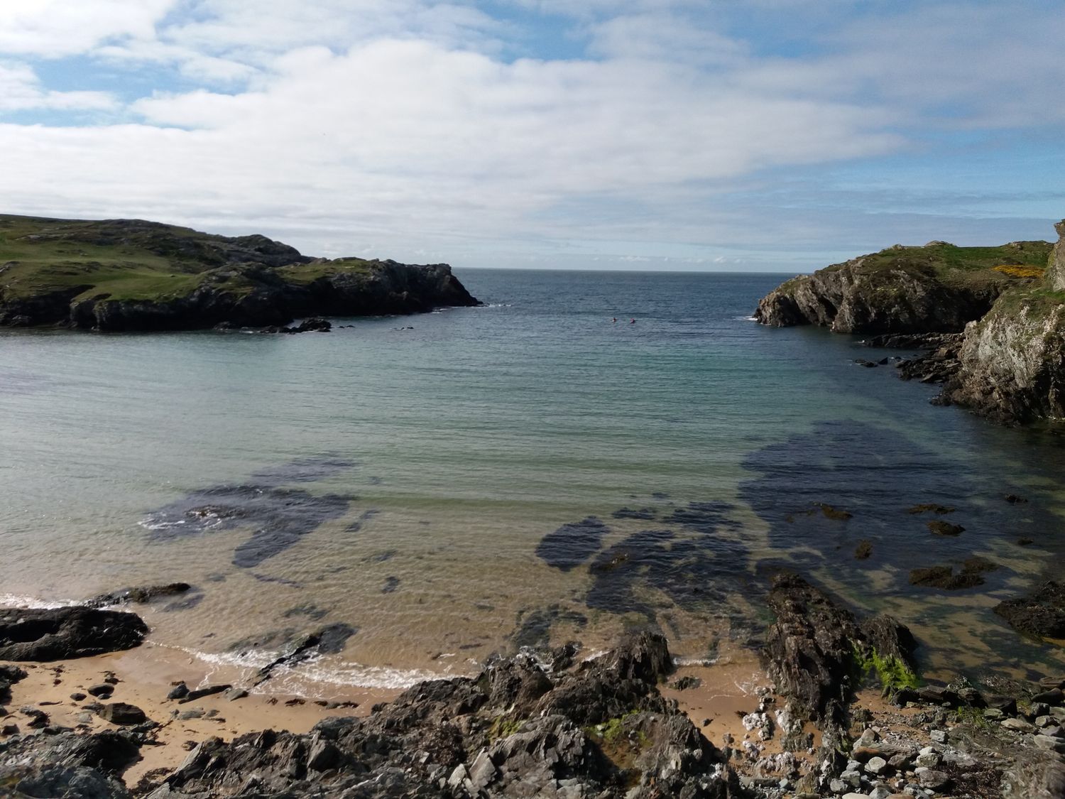 A rocky beach with clear water and grassy cliffs surrounding a small bay at Cysgod y Bugail in Holyhead