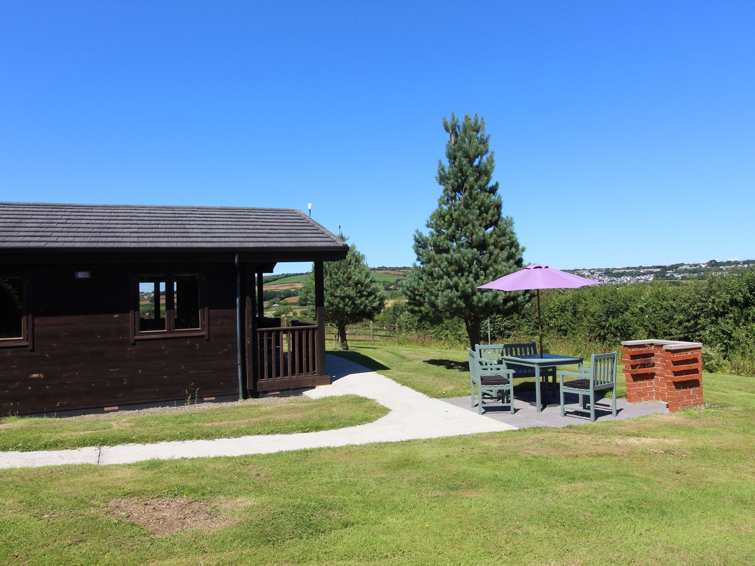 An outdoor seating area with chairs a table and umbrella next to a barbecue with a wooden cabin and trees in the background at Willow Lodge in Lanivet near Bodmin