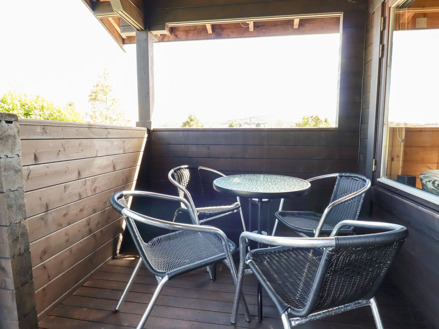 A covered wooden balcony with a round glass table and four black metal chairs at Hazel Lodge in Lanivet near Bodmin