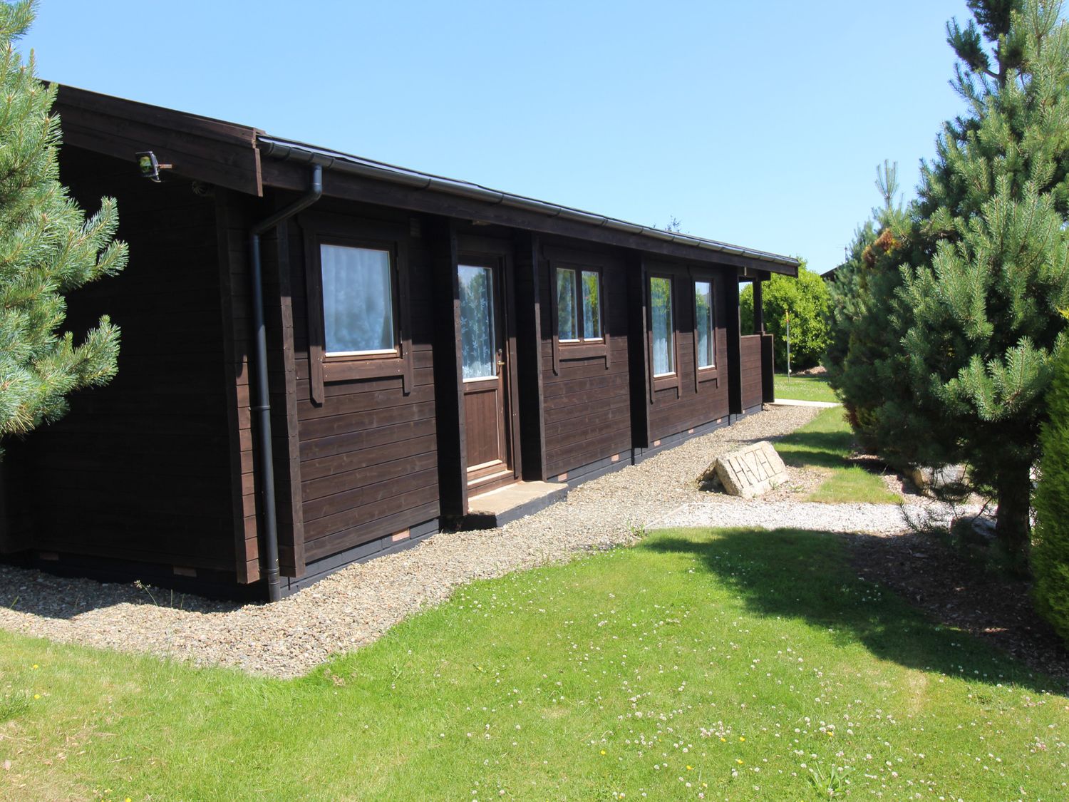 A dark wooden cabin with windows and a door surrounded by grass and trees at Hazel Lodge in Lanivet near Bodmin