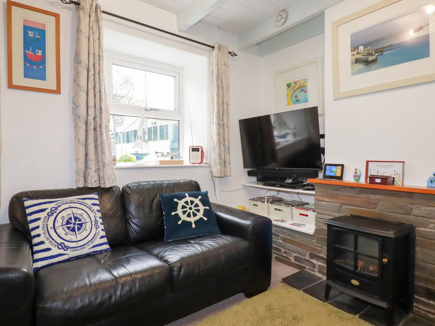 A living room with a black leather sofa two nautical-themed cushions a window with curtains a television on a white shelf a small stove and framed pictures on the walls at Daisy Cottage in Perranporth