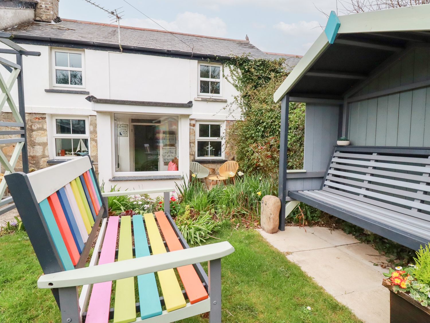 A garden area with a colorful bench and a covered gray bench in front of a white house with windows at Daisy Cottage in Perranporth