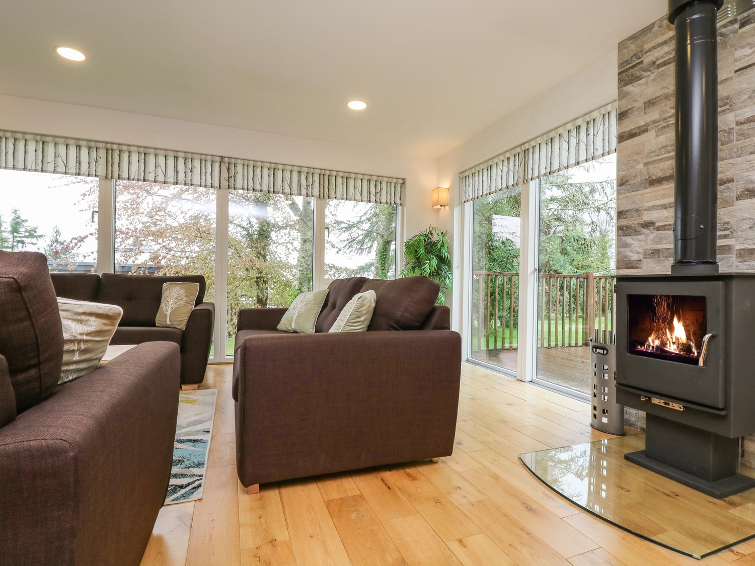 A living room with brown sofas a wood burning stove and large windows overlooking trees at Elm in Truro