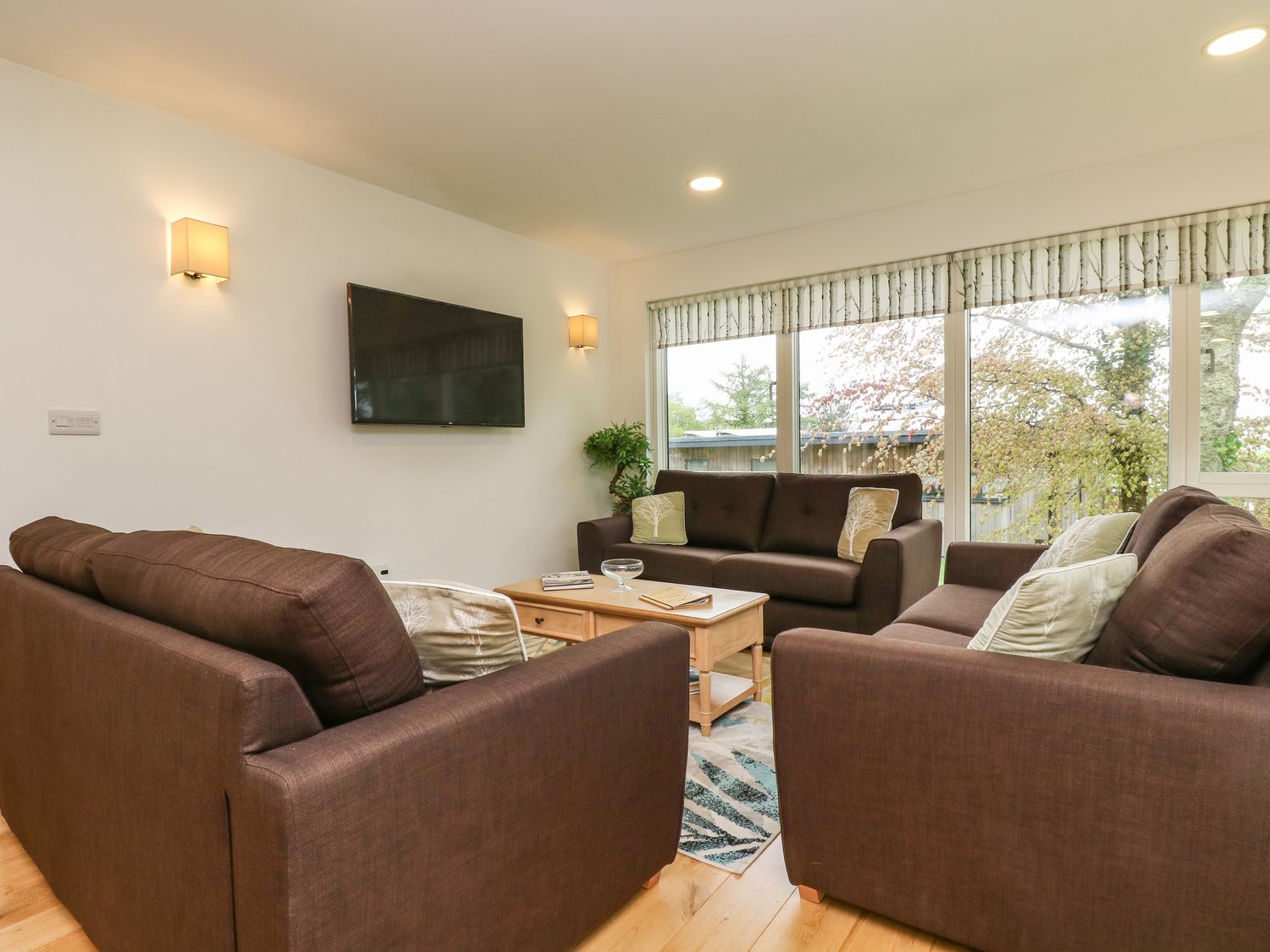 A living room with three brown sofas a wooden coffee table a wall-mounted television and large windows with patterned blinds at Elm in Truro