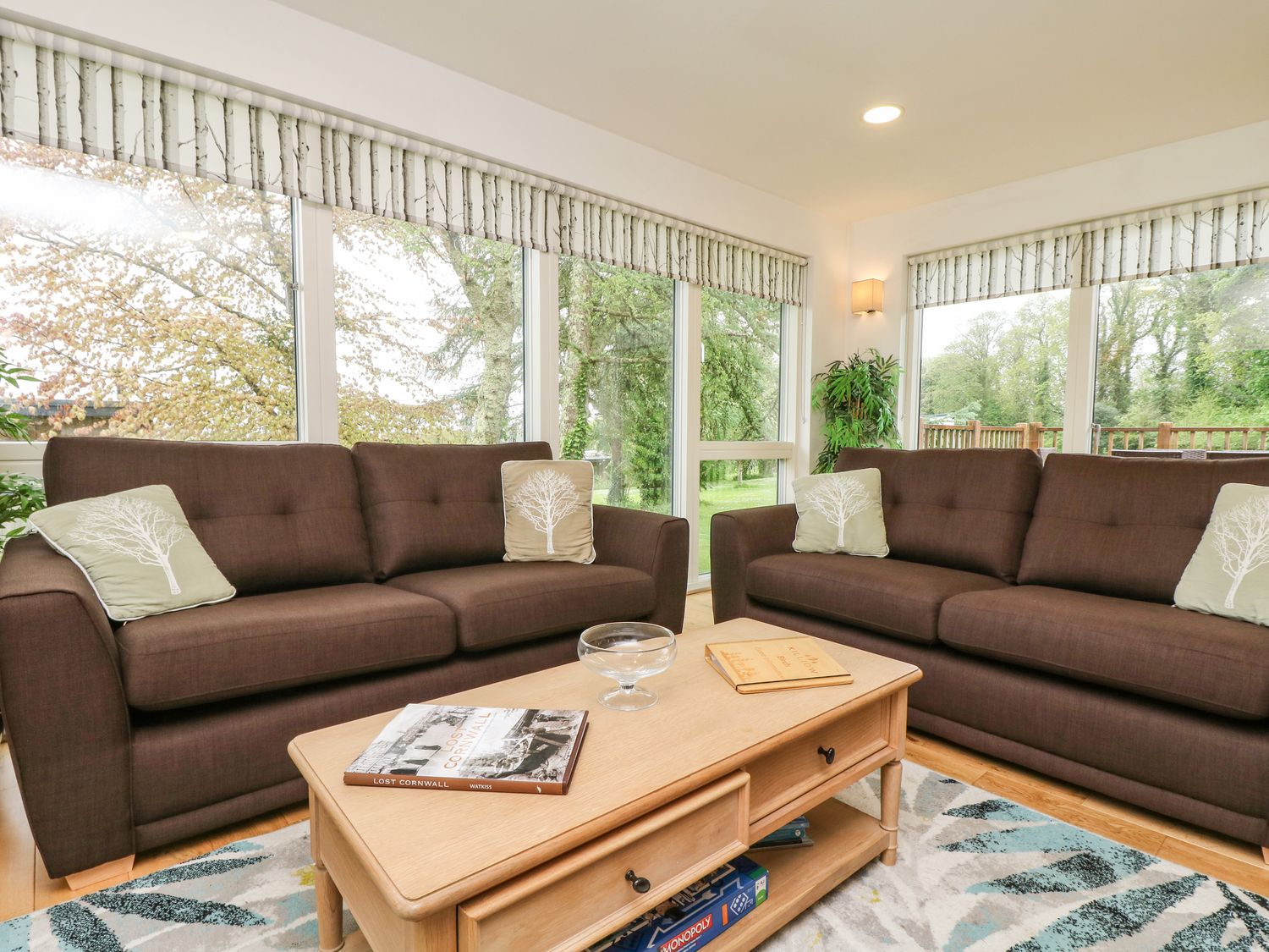 A living room with two brown sofas, green pillows with tree designs, a wooden coffee table with books and a glass bowl, and large windows with blinds at Elm in Truro