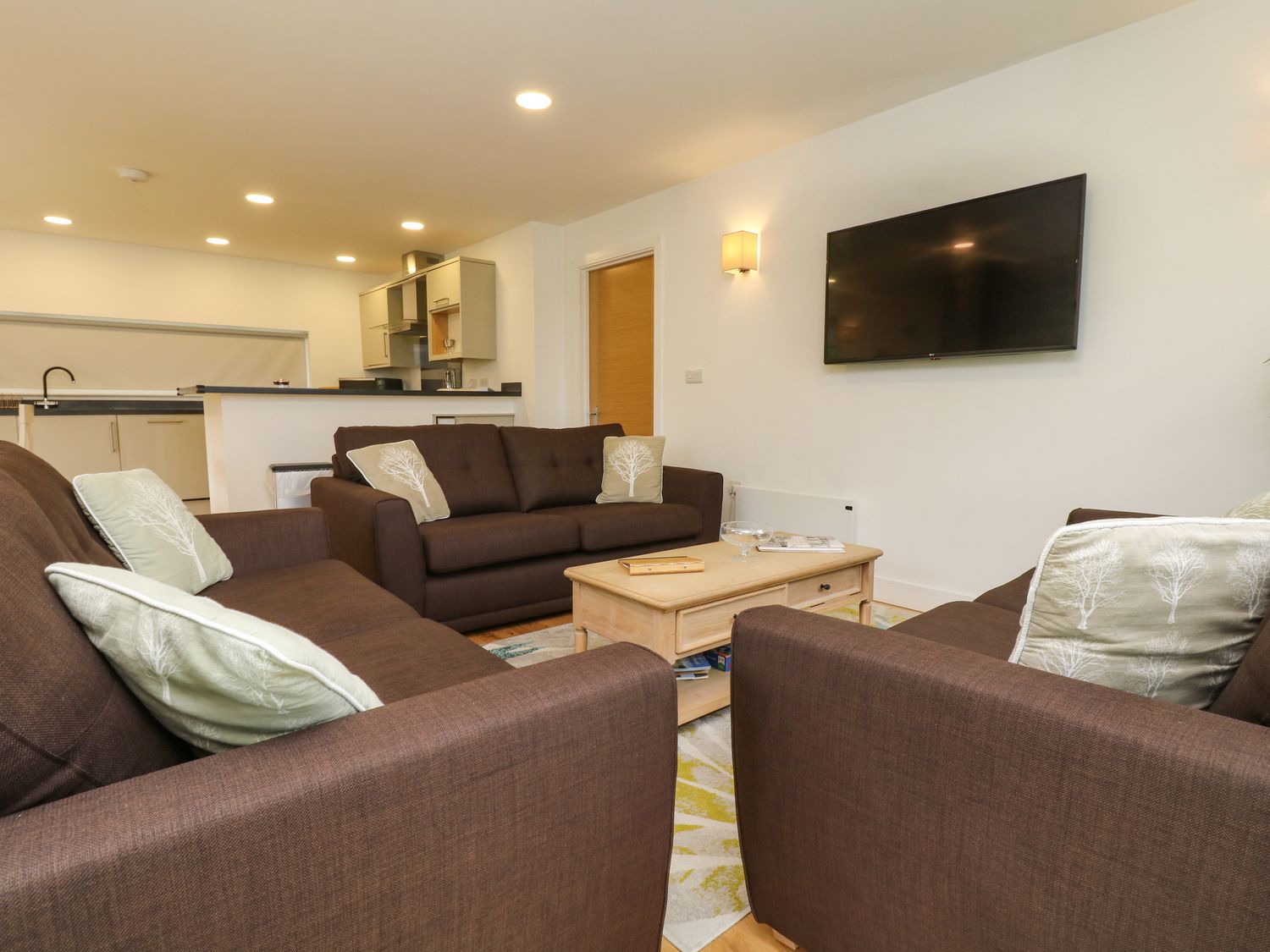 A living room with three brown sofas and cushions around a wooden coffee table with a TV mounted on the wall at Elm in Truro