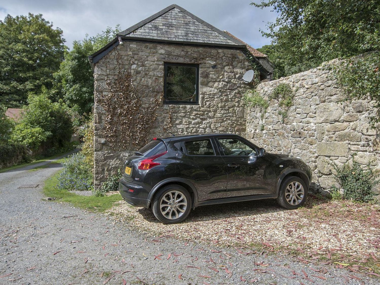 A black car parked beside a stone building and stone wall on a gravel surface with trees in the background at Water Barn in Manaton near Lustleigh