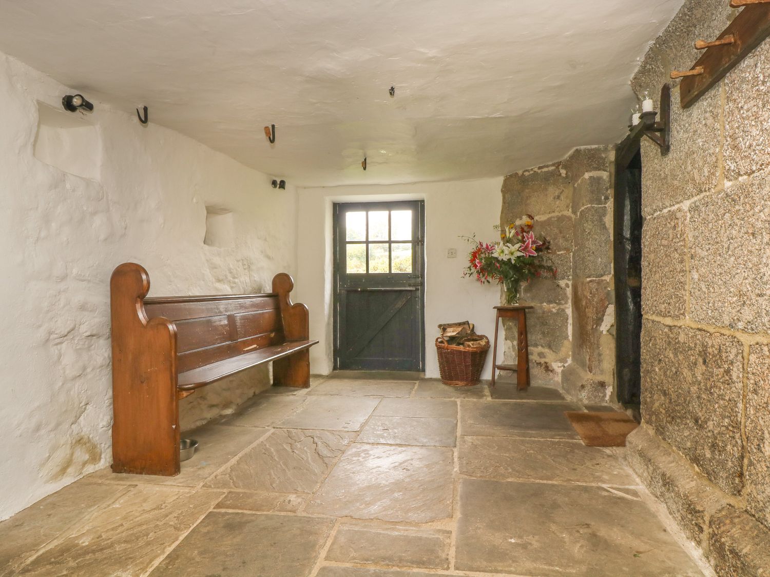 A room with a wooden bench a door a basket with firewood a small table with flowers and stone walls at Hole Farm in Chagford