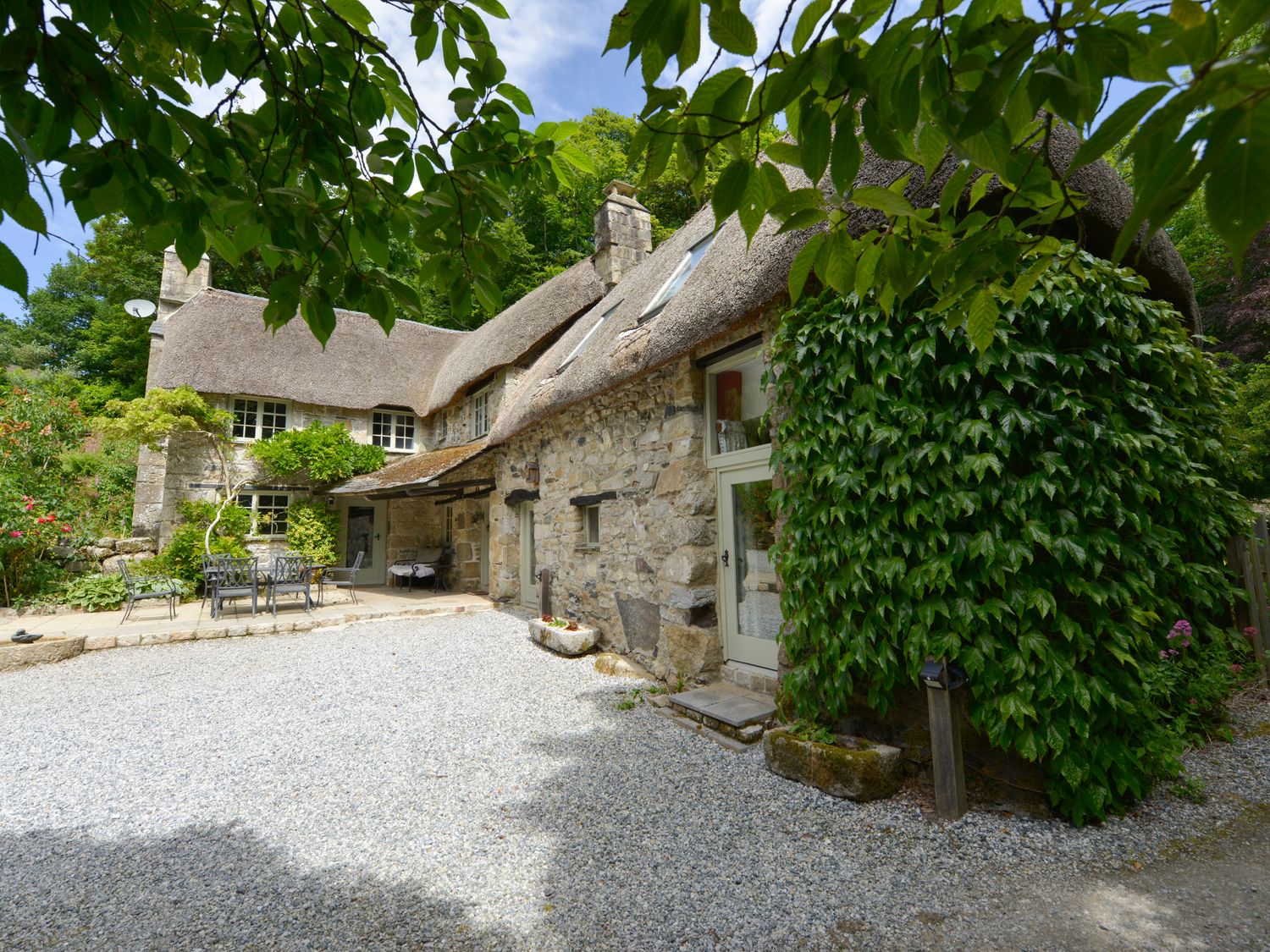 A stone cottage with a thatched roof and outdoor metal table and chairs surrounded by greenery at Bagtor Mill in Ilsington near Haytor Vale