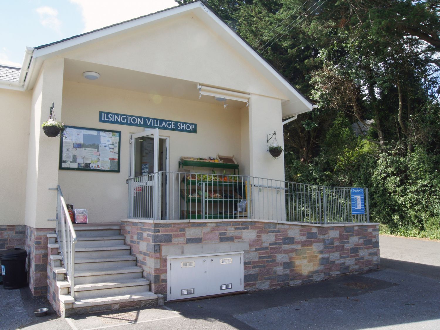 The entrance of Ilsington Village Shop with steps, railing, a doorway, hanging baskets, and a notice board at Ilsington Village Shop