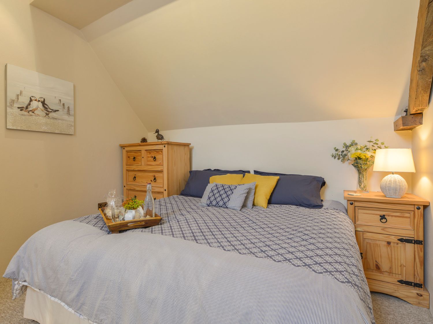 A bedroom with a bed with patterned bedding two wooden bedside tables a lamp flowers and a painting on the wall at The Bolthole in Throwleigh near Chagford