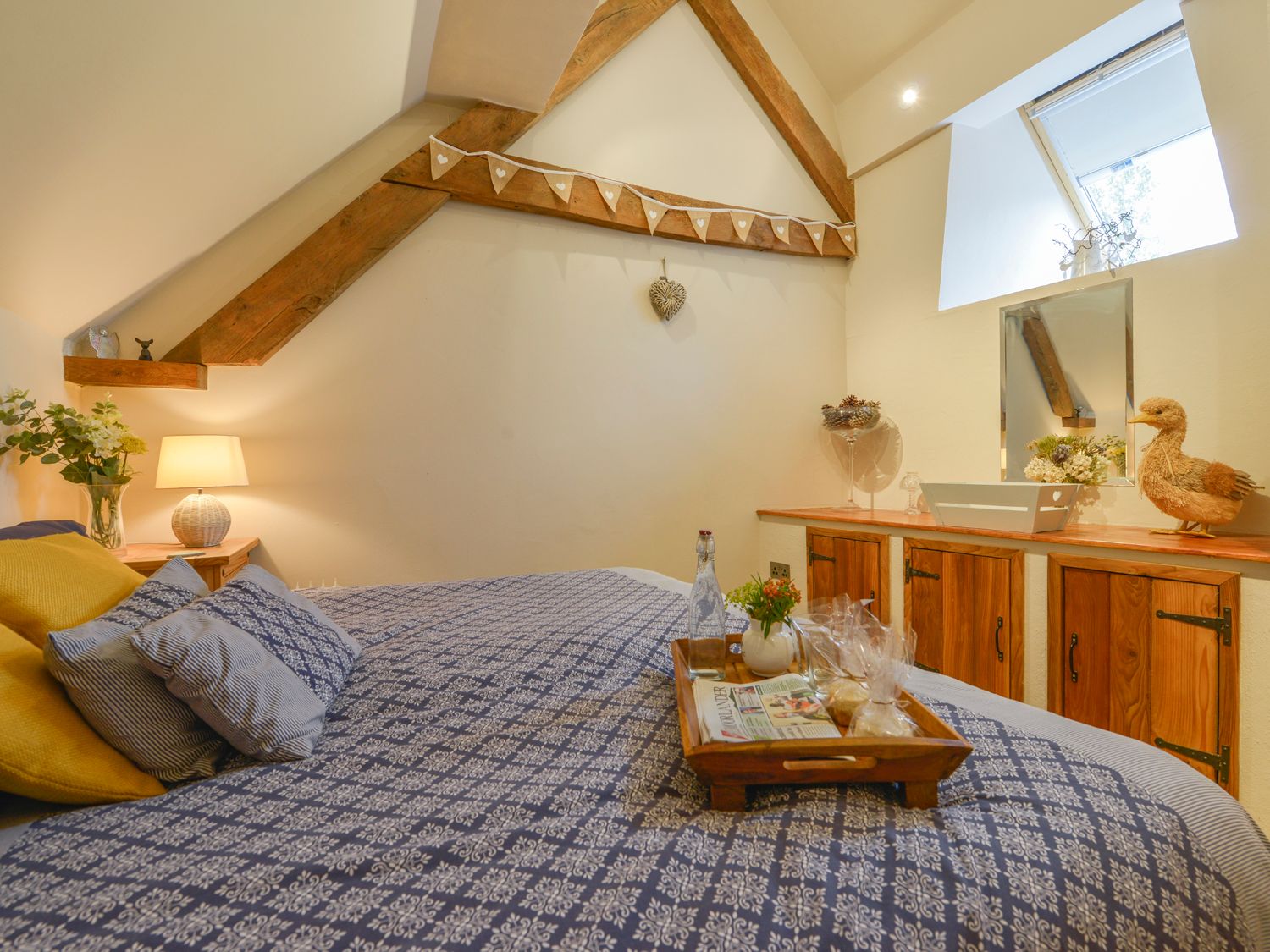 A bedroom with a bed covered in patterned bedding and cushions a tray with a bottle and snacks a wooden cabinet a small window and decorative items at The Bolthole in Throwleigh near Chagford