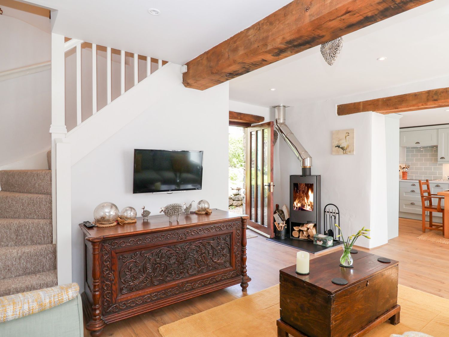 A living room with a television mounted above a carved wooden cabinet a wood-burning stove and a wooden chest table at The Bolthole in Throwleigh near Chagford