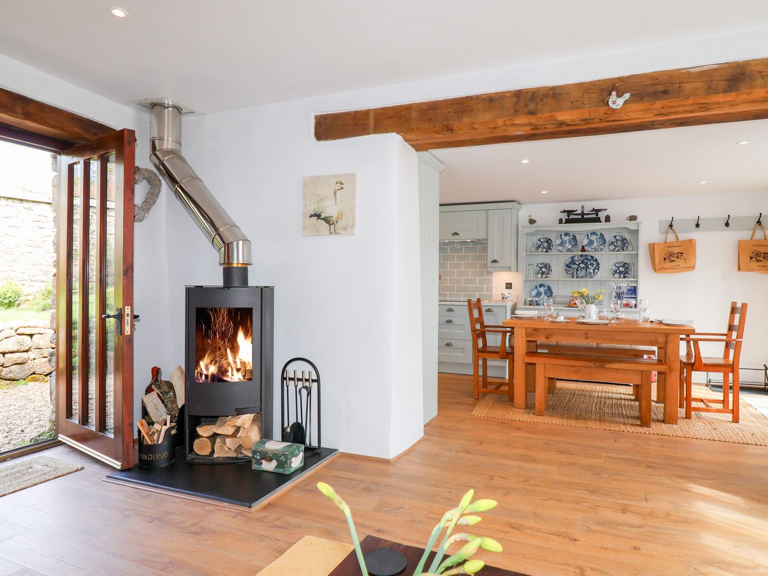 A room with a wood-burning stove and wood storage next to an open door and a dining table with chairs and a bench at The Bolthole in Throwleigh near Chagford